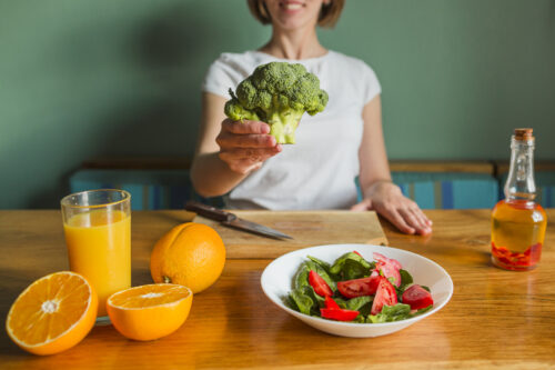 Mulher apresentando um brócolis como símbolo de alimentação saudável. A mesa está repleta de opções nutritivas, como suco de laranja, laranjas frescas, salada de espinafre e tomate, além de uma garrafa de azeite.