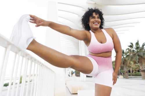 Essa imagem mostra uma mulher sorridente realizando um exercício de alongamento, representando disposição e bem-estar durante a prática de atividades físicas ao ar livre.