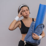 Mulher confusa e cansada segurando mochila com colchonete de yoga.