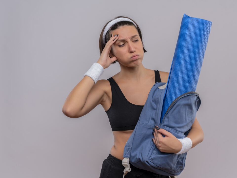 Mulher confusa e cansada segurando mochila com colchonete de yoga.