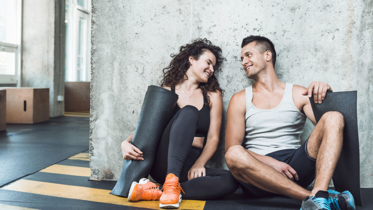 Casal aproveita momento de descontração na academia após um treino em dupla, reforçando os benefícios da parceria nos exercícios.