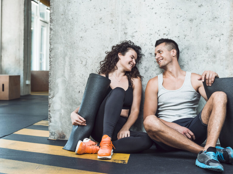 Casal aproveita momento de descontração na academia após um treino em dupla, reforçando os benefícios da parceria nos exercícios.